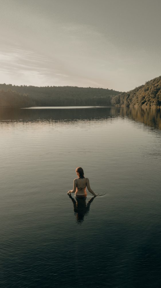 image description: a woman slowly wades into a body of water nestled with forests ahead of her