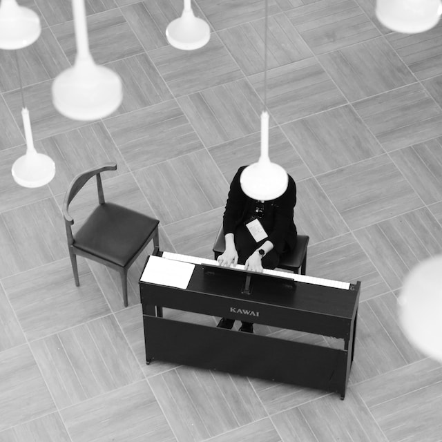 Image description: Black and white photo of an obscured person playing the piano with no audience, an empty chair sits beside them