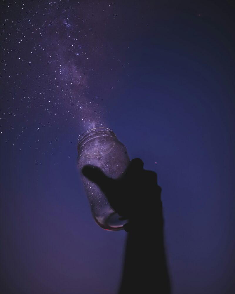 A hand holds a mason jar up to the sky to catch the stars